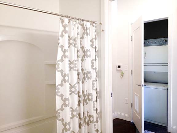A white bathroom with a shower curtain and a doorway leading to a kitchen.